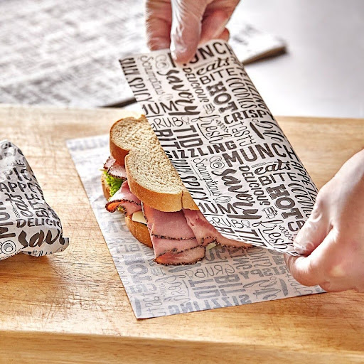 A hand wrapping a fresh deli sandwich with custom printed deli paper featuring restaurant typography.