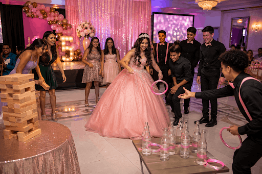 Guests and a young woman in a pink ballgown enjoying a giant Jenga game at a Quinceanera celebration.