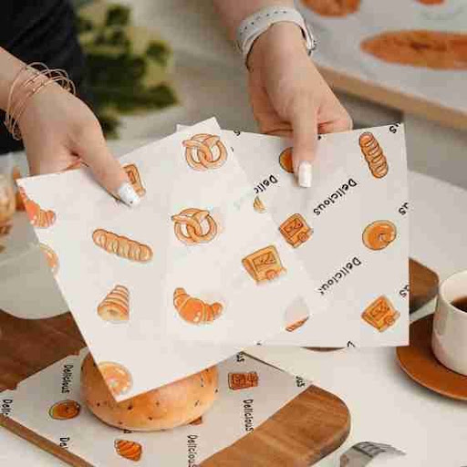 Hands placing custom printed greaseproof paper wraps with bakery designs over bread and pastries.