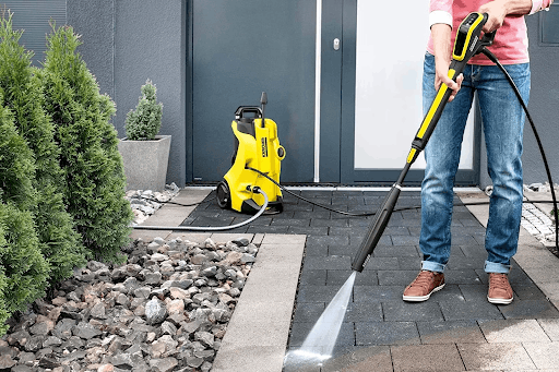 A yellow electric pressure washer spraying water on a stone paved patio next to green bushes.